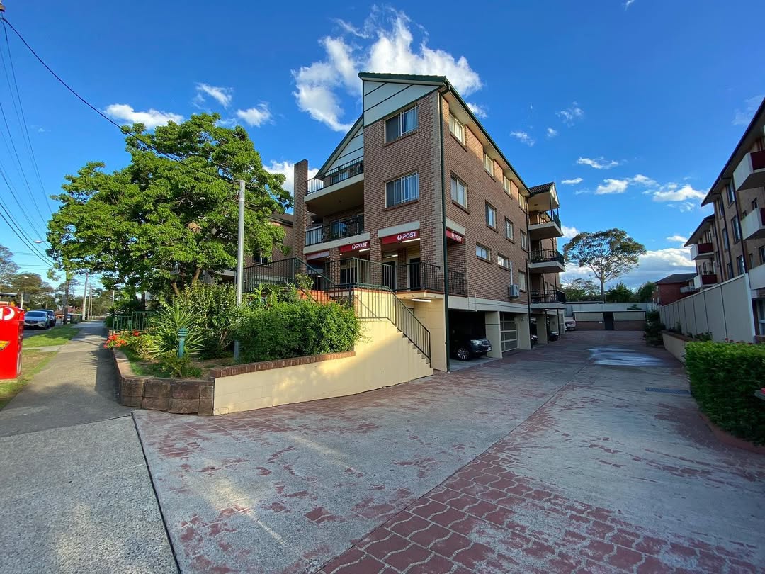 Three-storey brick apartment complex with balconies, driveway parking, and landscaped surroundings on a sunny day